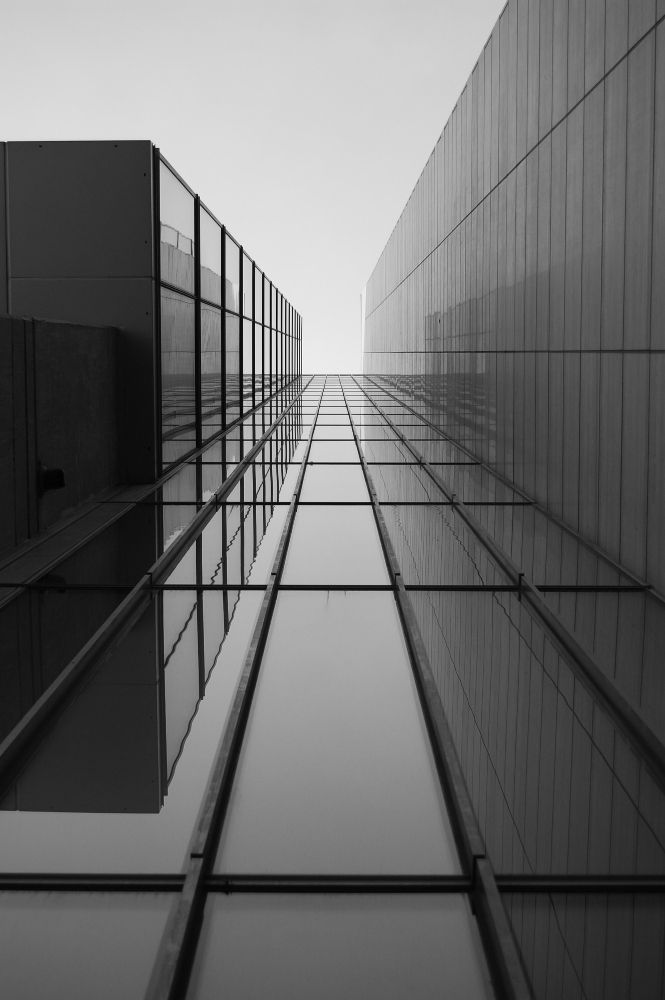 Afbeelding: Een zwart-wit foto die recht omhoog kijkt tussen twee moderne glazen wanden. De verticale lijnen convergeren extreem naar het midden toe tegen een lichte lucht