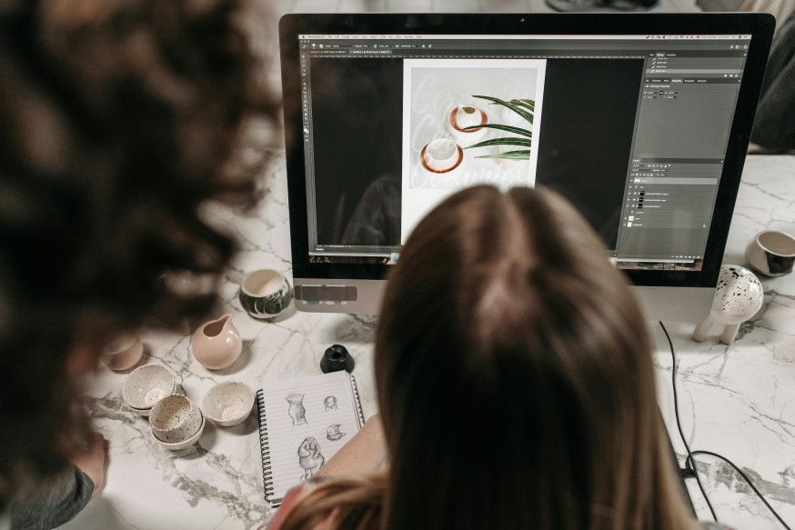 Afbeelding: Bovenaanzicht van iemand die schetsen maakt op papier terwijl ze op een computerscherm kijkt naar creatieve ontwerpen