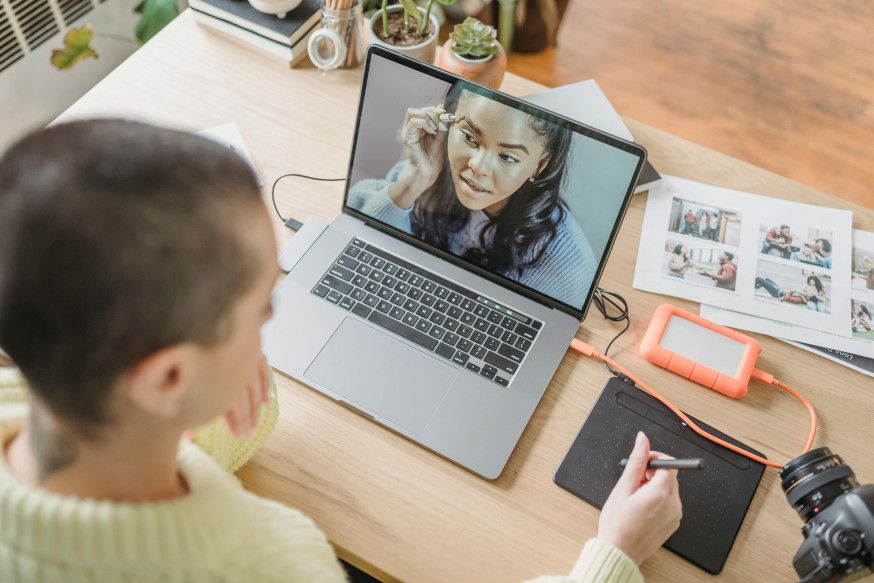 Afbeelding: Creatief persoon aan een bureau werkt met een tekentablet en laptop, waarop een digitaal (AI-)portret met make-up detail zichtbaar is, omringd door referentiefoto's
