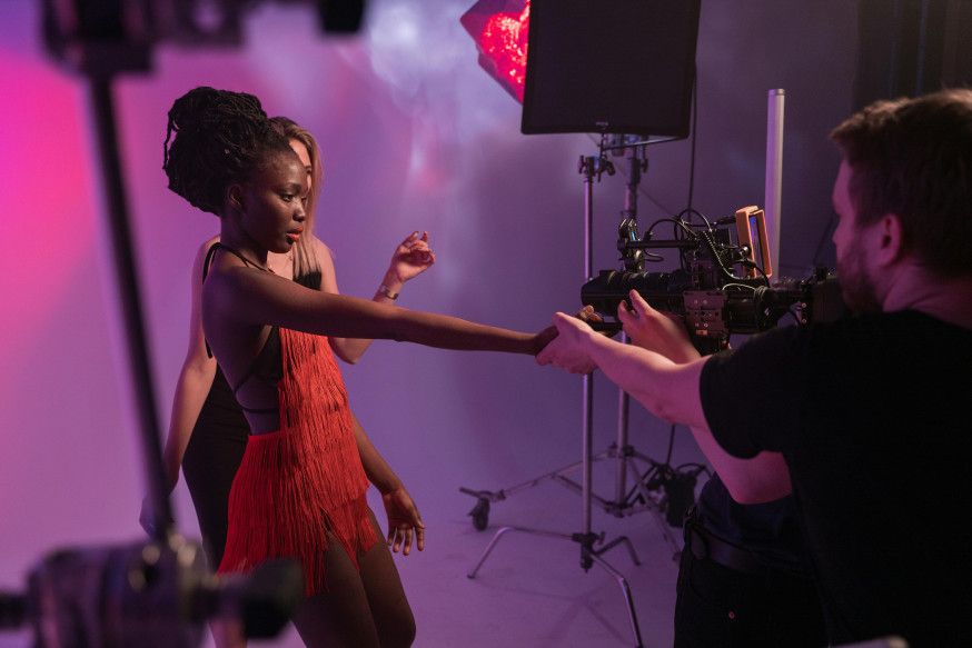 Afbeelding: Fotograaf op een filmset geeft aanwijzingen aan een model in een rode jurk, met professionele studiolampen en apparatuur