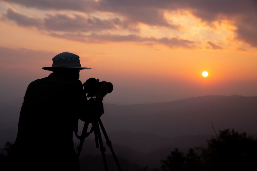 Afbeelding: Silhouet van een fotograaf die achter een camera op statief staat tijdens zonsondergang