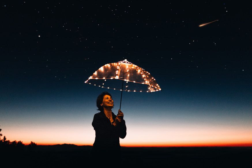 Afbeelding: Vrouw staat in het donker onder een verlichte paraplu met een heldere sterrenhemel op de achtergrond, als voorbeeld van hoe mooi avond fotografie kan zijn