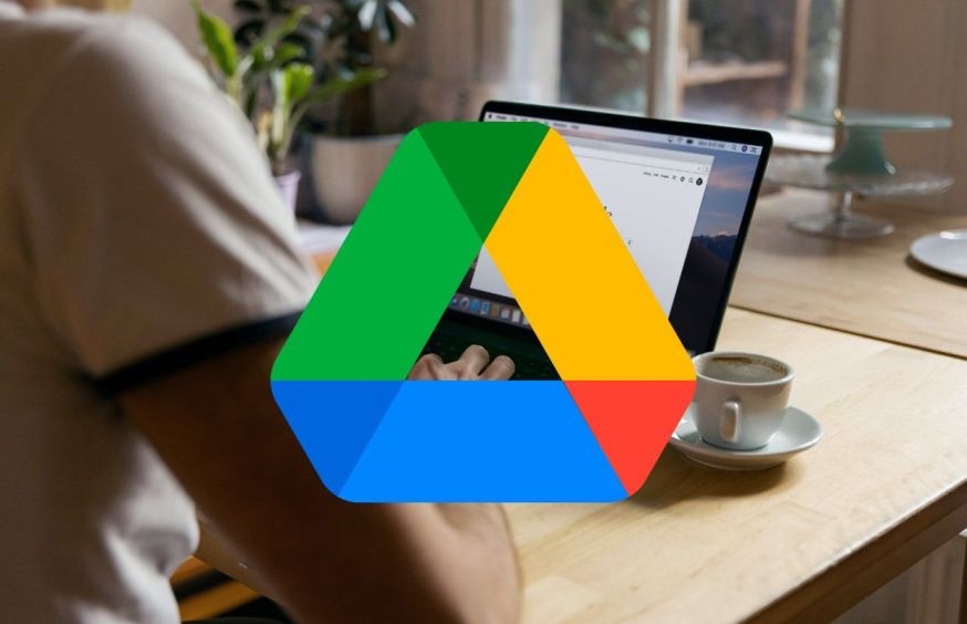Afbeelding: Het bekende groen-geel-blauw-rode Google Drive-logo groot in beeld gebracht, met op de achtergrond een wazige setting van een persoon die aan een houten tafel op een laptop werkt naast een kop koffie