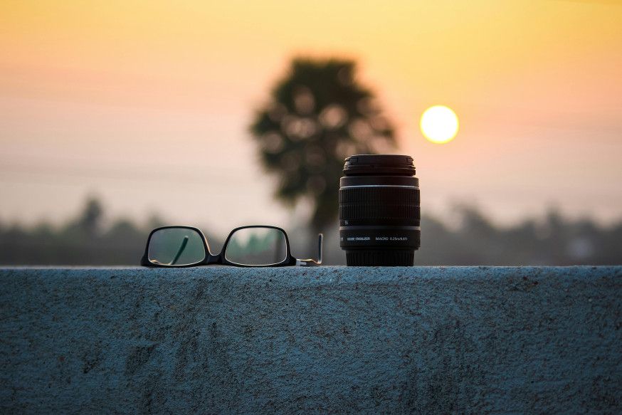 Afbeelding: Een cameralens en een bril liggen op een muurtje tijdens een zonsondergang